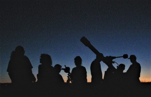 Silhouettes of people observing the night sky with telescopes against a twilight backdrop.