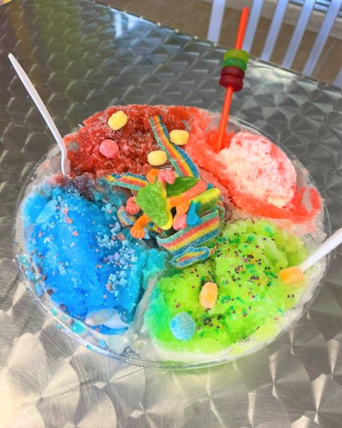 A colorful bowl of shaved ice with various flavors, topped with candy and a straw, on a metallic surface.