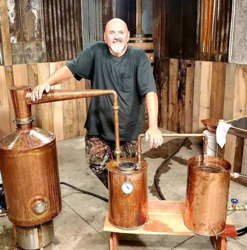 A man stands beside a copper distillation setup in a rustic workshop, smiling at the camera.