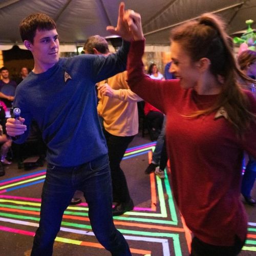 A young man and woman dance together under a tent, wearing matching colorful shirts with a sci-fi theme.