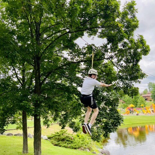 A person in a helmet zip lines between trees over a grassy area near a pond, with playground equipment in the background.