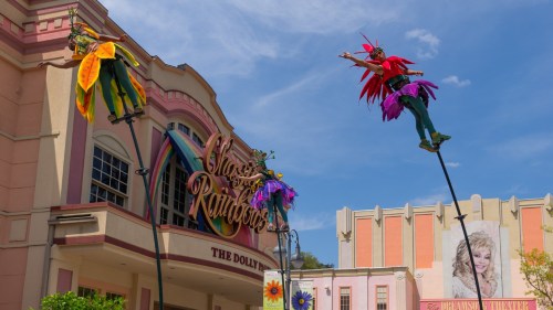 Colorful performers on stilts entertain in front of a vibrant building at Dollywood's "Chasing Rainbows" attraction.