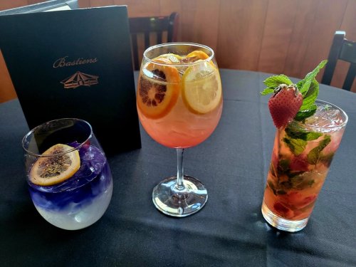 Three colorful cocktails on a black table: a blue drink with lemon, a pink drink with citrus, and a minty strawberry drink.
