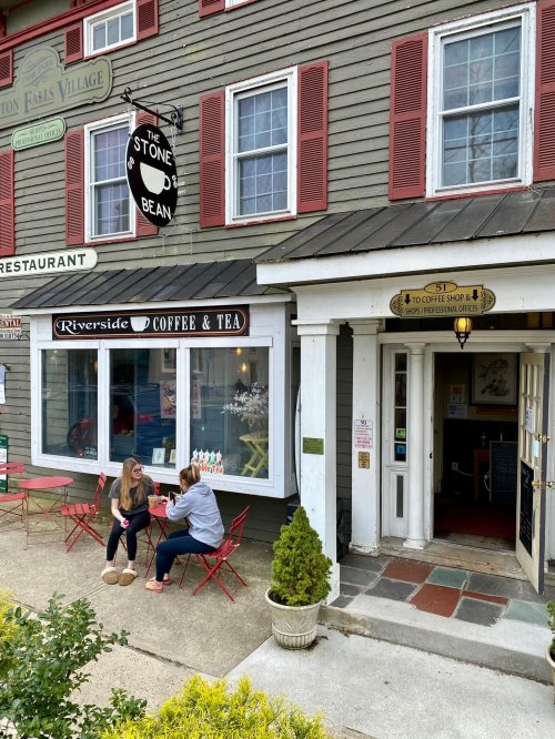 Two women sit at a small table outside a quaint coffee shop with red chairs and a sign reading "The Stone Bean."