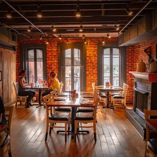 Cozy restaurant interior with wooden tables, brick walls, and large windows, featuring two diners engaged in conversation.