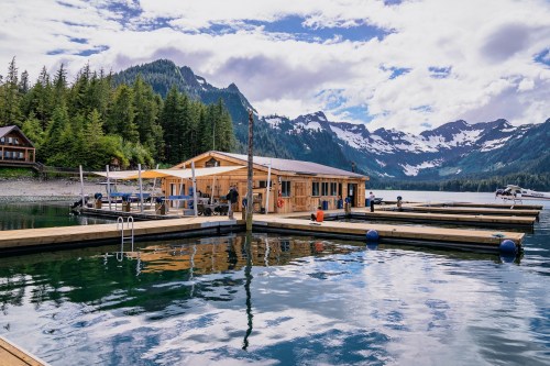 A scenic lakeside view featuring a wooden dock, a cabin, and mountains in the background under a partly cloudy sky.