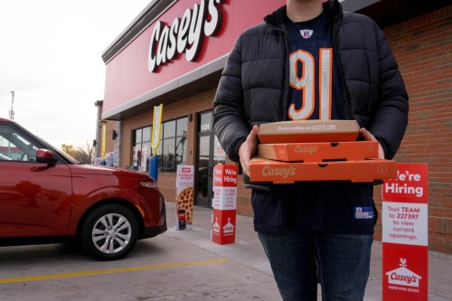 A person in a sports jersey holds multiple pizza boxes outside a Casey's store, with a car parked nearby.