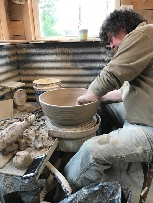 A potter shapes a large bowl on a wheel, surrounded by clay and tools in a sunlit studio.