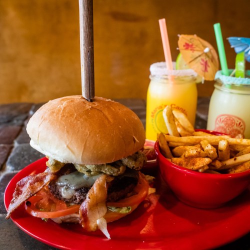 A juicy burger topped with bacon and pickles, served with fries in a red bowl, alongside colorful drinks with umbrellas.
