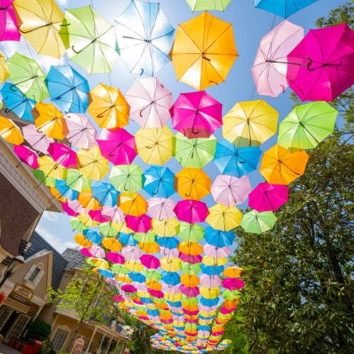 Colorful umbrellas in vibrant shades hang overhead, creating a cheerful canopy against a bright blue sky.