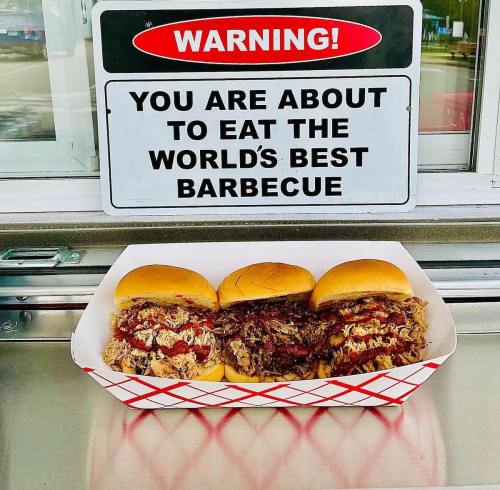 A tray with three barbecue sandwiches in front of a sign warning about the world's best barbecue.