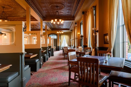 Elegant restaurant interior with wooden tables, plush seating, and ornate ceiling, featuring warm lighting and large windows.