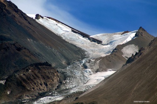 A mountainous landscape featuring a glacier flowing down a rocky slope under a clear blue sky.