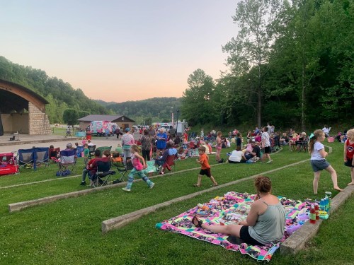 A lively outdoor gathering at dusk, with people sitting on blankets and chairs, enjoying a community event in a green park.