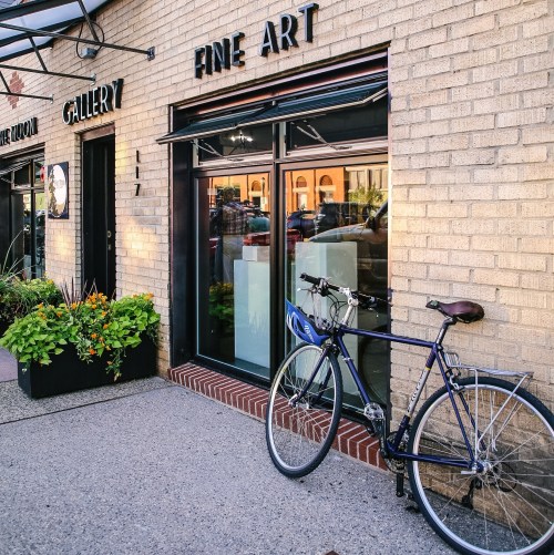 A blue bicycle leans against a brick wall outside a fine art gallery with large windows and colorful flowers.