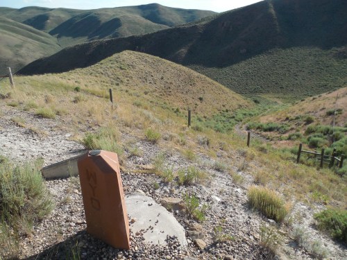 A weathered marker labeled "WY" stands on a gravel path, surrounded by rolling green hills and sparse vegetation.