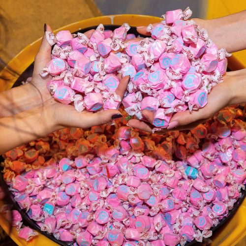 Two hands hold a handful of colorful pink and blue candies above a large bowl filled with more candies.
