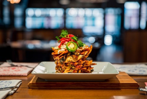 A stacked plate of nachos topped with jalapeños, red peppers, and fresh herbs, served on a wooden board.
