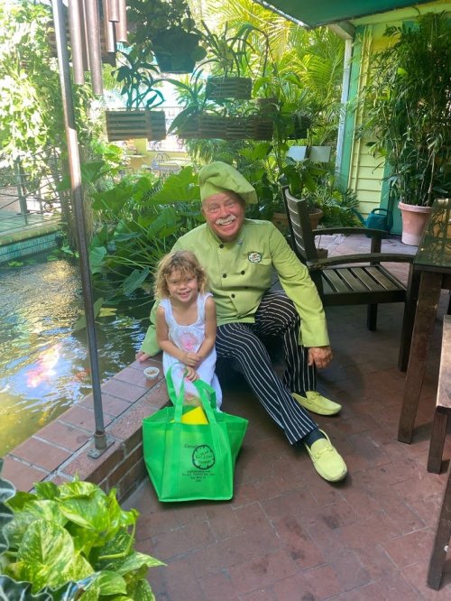 A smiling chef in a green outfit sits beside a young girl with curly hair, surrounded by lush greenery and a pond.