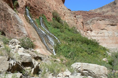 A waterfall cascading down rocky cliffs, surrounded by lush greenery and rugged terrain under a clear blue sky.