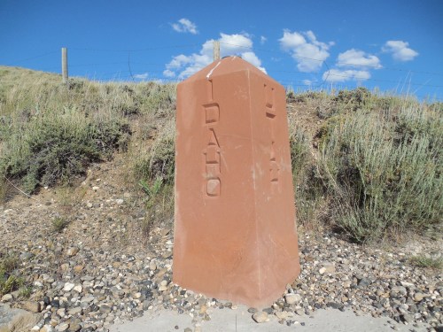 A stone marker with "IDAHO" engraved, surrounded by rocky terrain and sparse vegetation under a blue sky.