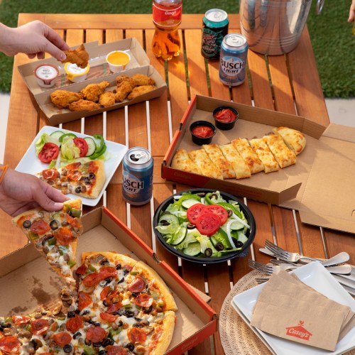 A table spread with pizza, wings, salad, and drinks, set outdoors on a sunny day.