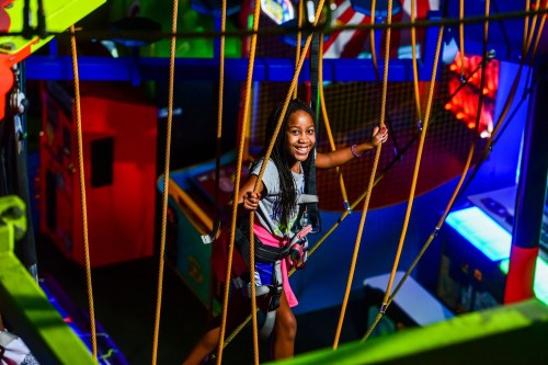 A smiling girl navigates a colorful indoor climbing structure, secured with a harness and ropes.