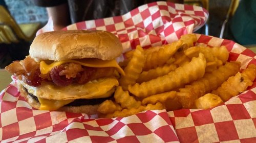 A burger with cheese, bacon, and a fried egg, served with crinkle-cut fries in a red-checkered basket.