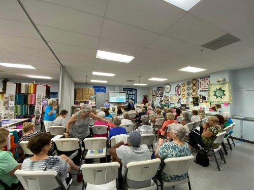 A large group of people seated in a classroom, watching a presentation with quilts displayed on the walls.