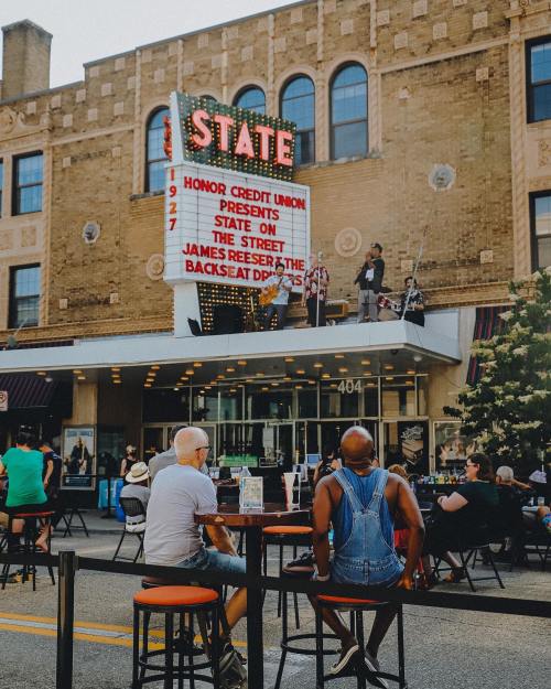 A crowd sits outside the State Theatre, enjoying a performance under a marquee that reads "State on the Street."