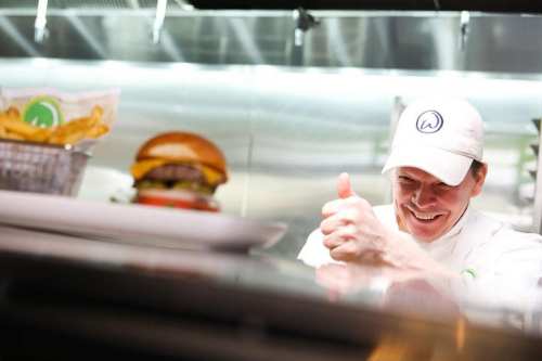 A chef gives a thumbs up in a kitchen, smiling at a burger and fries on the counter.