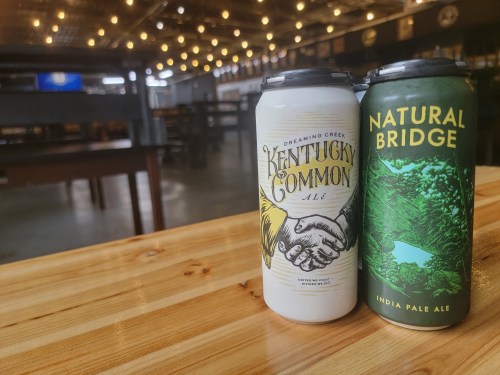 Two beer cans on a wooden table, with a blurred bar background and string lights overhead. One can is labeled "Kentucky Common" and the other "Natural Bridge."