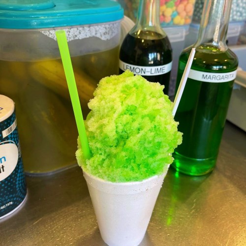 A vibrant green snow cone in a white cup, topped with two straws, with colorful syrups in the background.