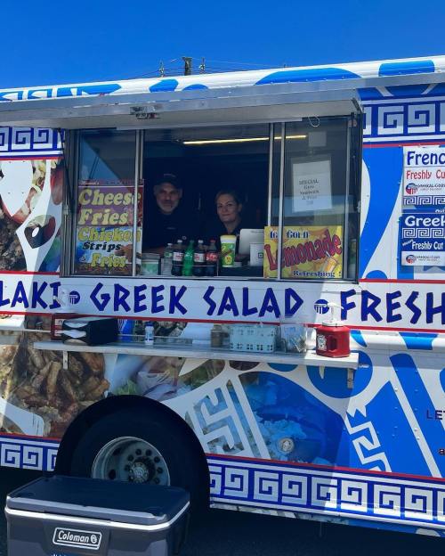 A colorful food truck with a Greek theme, featuring two staff members and a menu of salads, fries, and drinks.