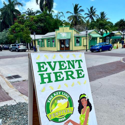 A sign promoting the Key Lime Festival in a vibrant, tropical setting with palm trees and colorful buildings.