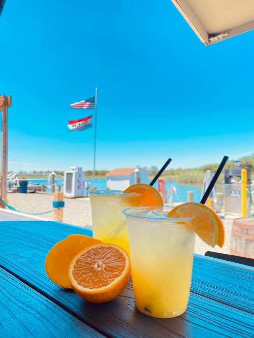 Two refreshing drinks with orange slices sit on a wooden table, with flags and a scenic waterfront in the background.