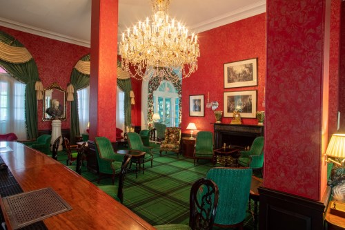 Elegant room with red walls, a chandelier, green chairs, and a fireplace, featuring classic decor and a patterned carpet.