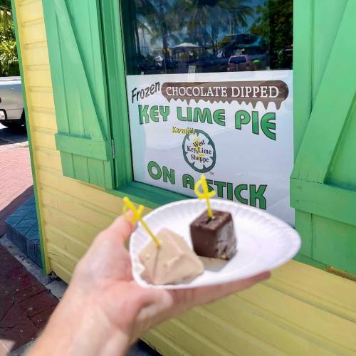 A hand holds a plate with chocolate-dipped key lime pie on a stick in front of a colorful shop window.
