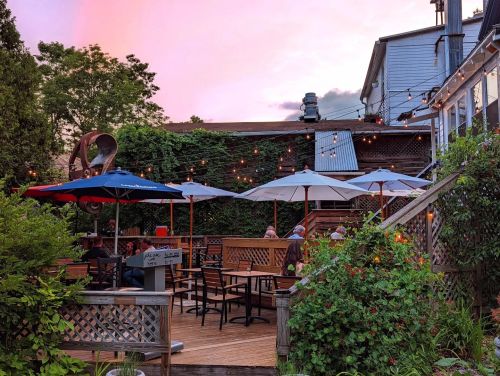 A cozy outdoor restaurant patio with umbrellas, string lights, and greenery under a colorful sunset sky.