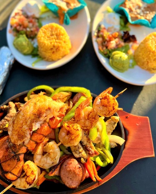 A sizzling platter of grilled seafood and vegetables, with two plates of rice, guacamole, and salsa in the background.