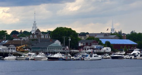 A scenic harbor with various boats docked, featuring buildings and church steeples in the background under a cloudy sky.