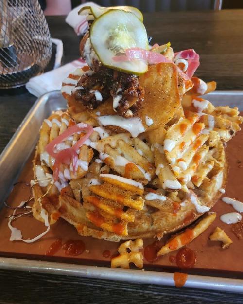 A towering stack of waffle fries topped with fried chicken, pickles, and drizzled with sauces on a metal tray.