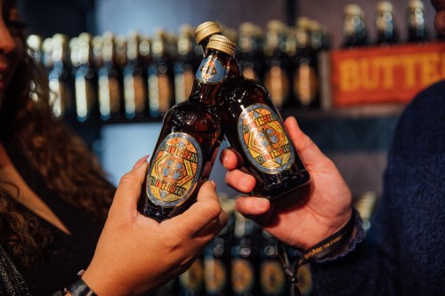 Two hands hold bottles of a dark beverage with decorative labels, set against a backdrop of more bottles on a shelf.