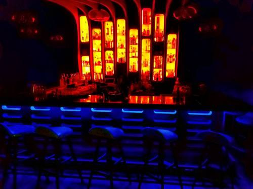 A dimly lit bar with a glowing blue counter and shelves of illuminated bottles in warm orange hues.