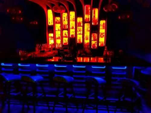 A dimly lit bar with a glowing blue counter and shelves of illuminated bottles in warm orange hues.