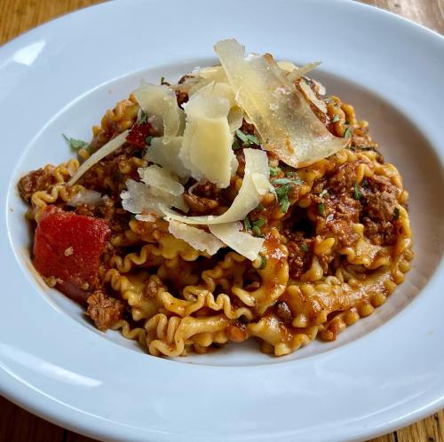 A plate of pasta with meat sauce, topped with shaved cheese and herbs, served in a white bowl.