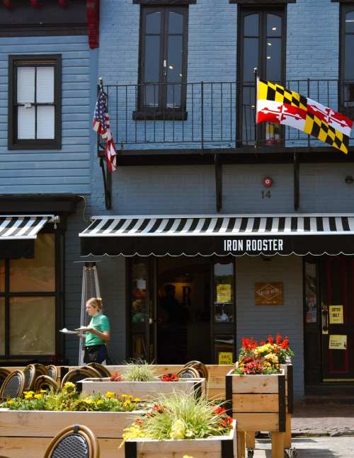 A woman carries a plate outside the Iron Rooster restaurant, with flags and colorful flowers in a vibrant outdoor setting.