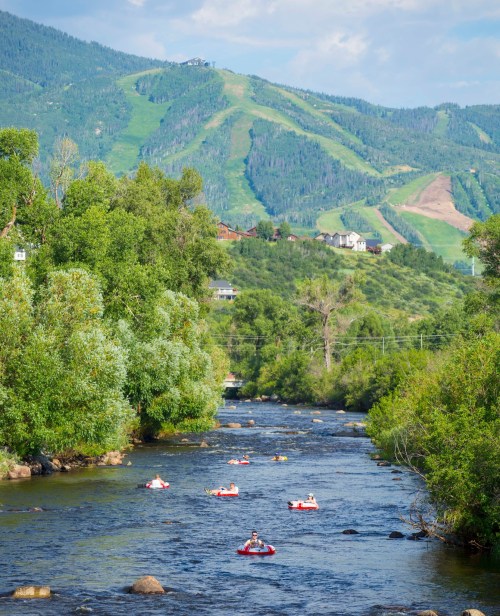 A scenic river with people floating on inner tubes, surrounded by lush greenery and mountains in the background.