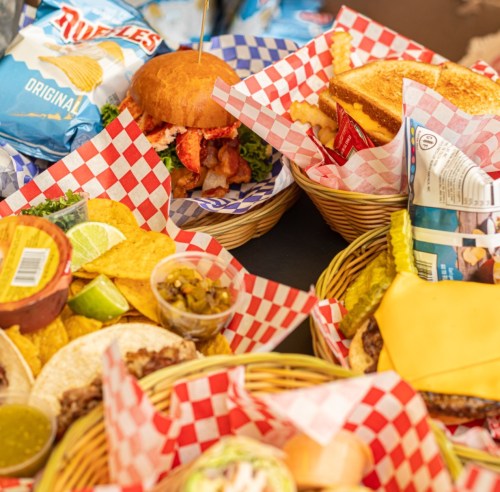 A variety of food in baskets, including a sandwich, tacos, chips, and dips, all served with colorful checkered liners.