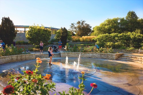 A sunny park scene with children playing in a fountain, surrounded by flowers and greenery. Families enjoy the outdoors.